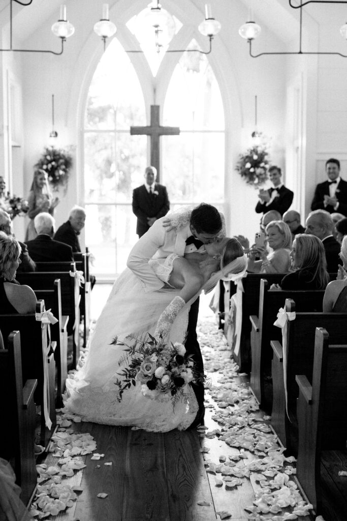 A groom in his white tuxedo dips and kisses his bride in the chapel aisle as guests watch during their montage palmetto bluff wedding ceremony