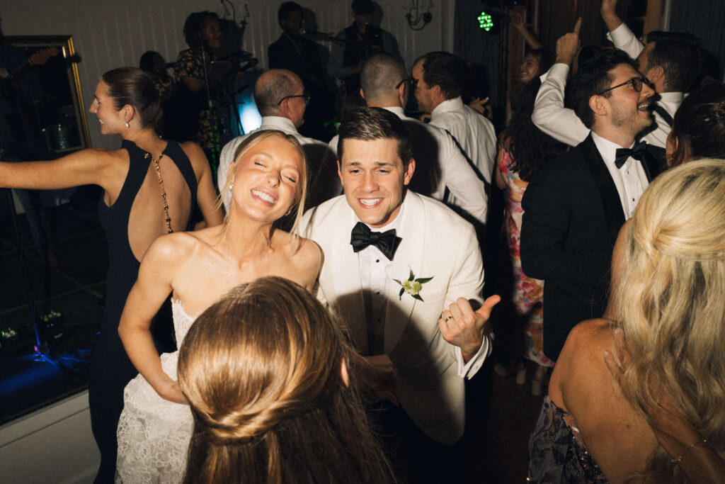A bride and groom laugh and dance with guests on the dance floor to their live band
