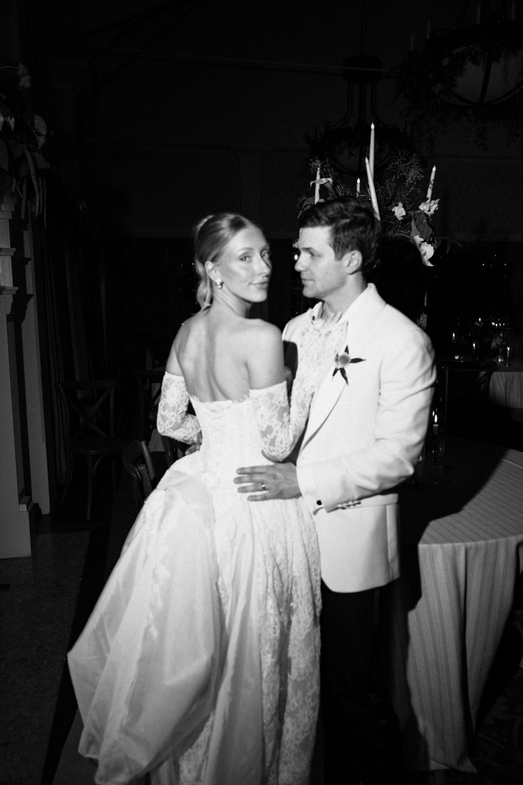 A bride smiles while cuddling her groom against a reception table in black and white