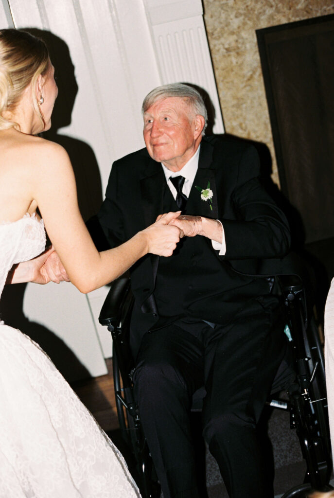 A bride leans in to chat with a smiling grandfather in a wheelchair at her reception