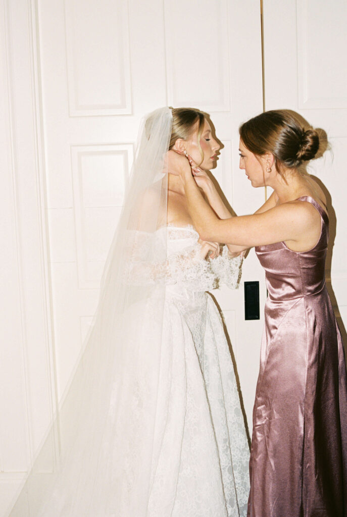 A bridesmaid in pink helps the bride put on earrings
