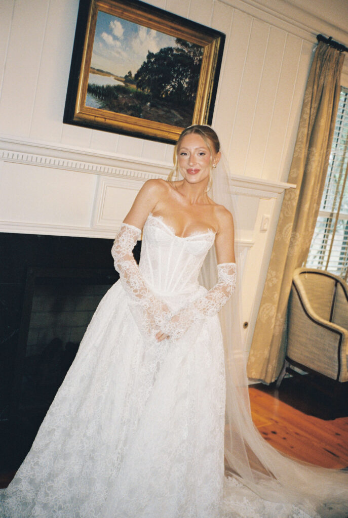 A bride smiles while standing by a fireplace in her gown and lace sleeves