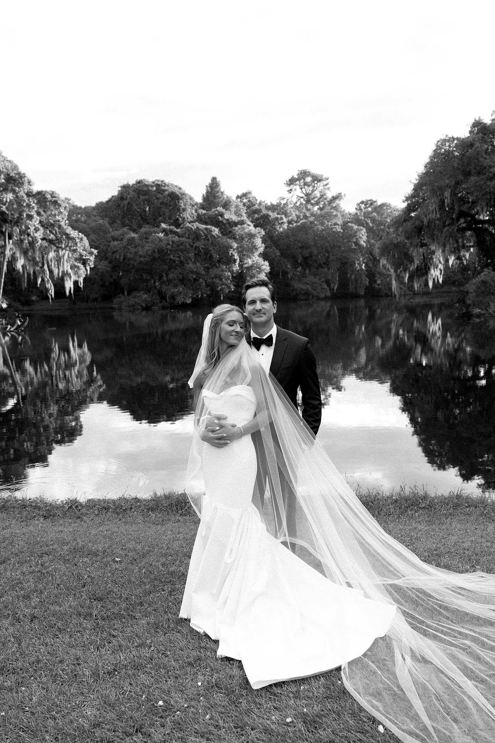 A bride and groom in black and white standing by the water snuggling and smiling at the legare waring house wedding venue